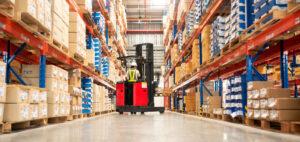 A worker in a forklift loading packed goods in a large distribution warehouse with high shelves